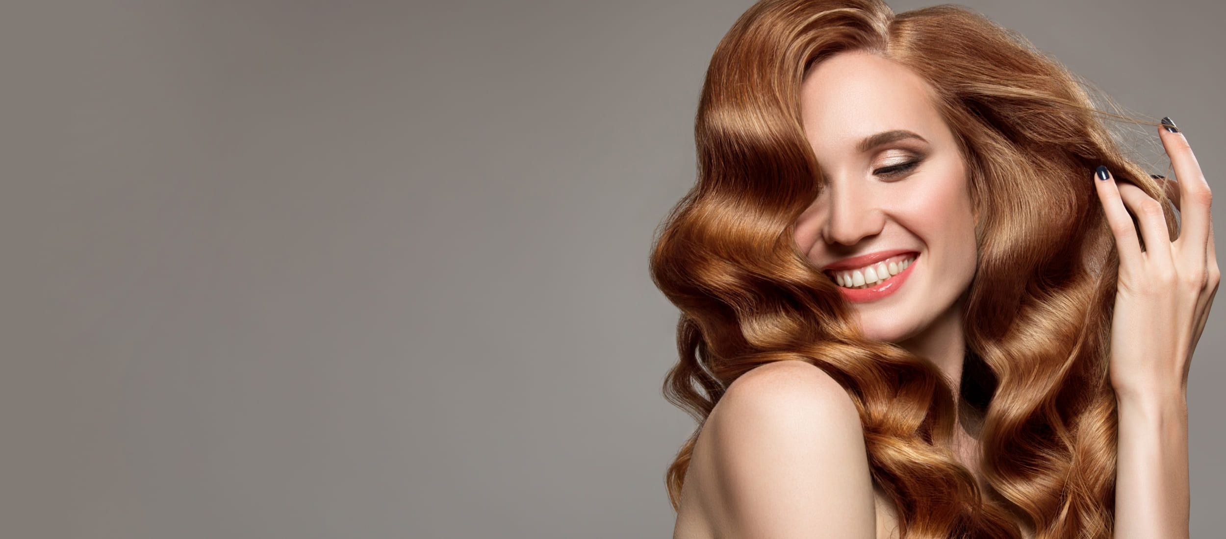 Frau mit langen, glänzenden, welligen Kupferhaaren lächelt und berührt sanft ihre Frisur - professionelle Haarpflege und Styling.